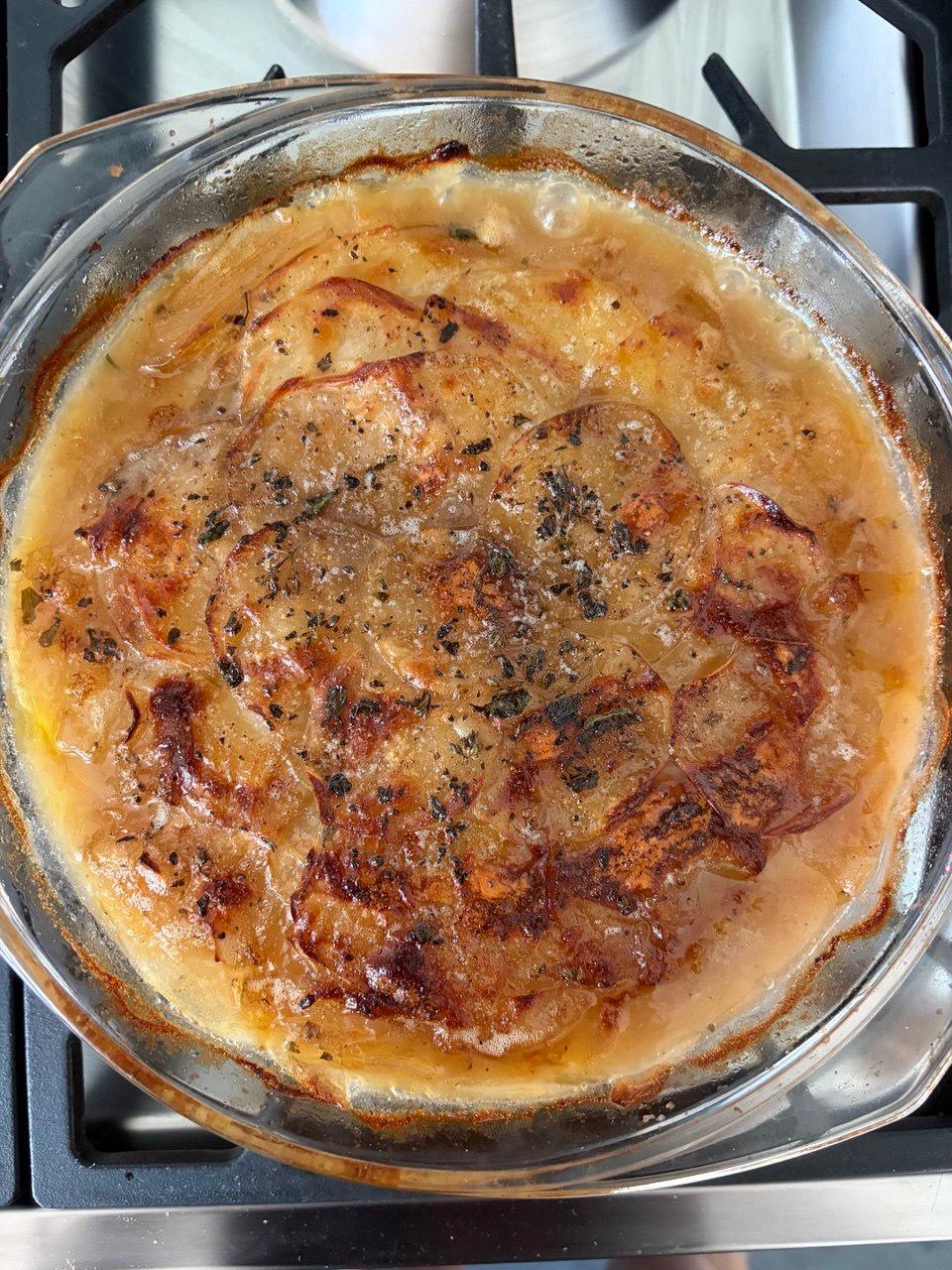 A round glass baking dish of boulangère potatoes, freshly out of the oven, with a deeply caramelised golden top speckled with dark herbs, sitting on a stainless steel gas hob.