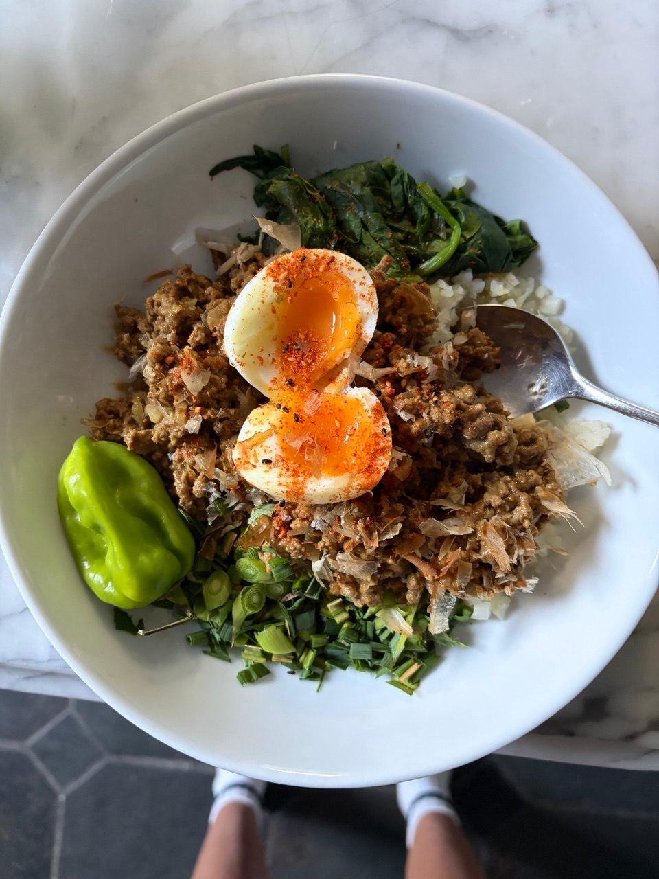 Overhead view of a white bowl containing Japanese curry mince over a bed of rice, topped with two halves of a soft-boiled egg dusted with togarashi, with wilted spinach, chopped spring onions and a whole green pepper arranged alongside, served with a spoon on a marble surface