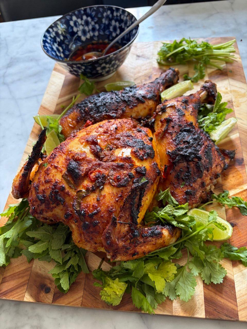 A whole spatchcocked chicken with deeply charred, rendang-red skin rests on a wooden chopping board surrounded by fresh coriander, mint and lime wedges, with a small blue patterned bowl of dipping sauce in the background.