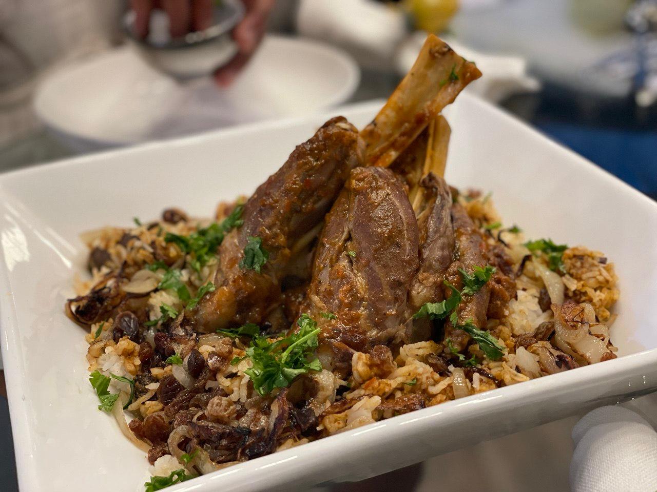 Two slow-braised lamb shanks with exposed bones standing upright, resting on a bed of spiced brown rice scattered with caramelised onions, raisins and toasted almonds, garnished with fresh coriander, served in a square white bowl on a restaurant table.