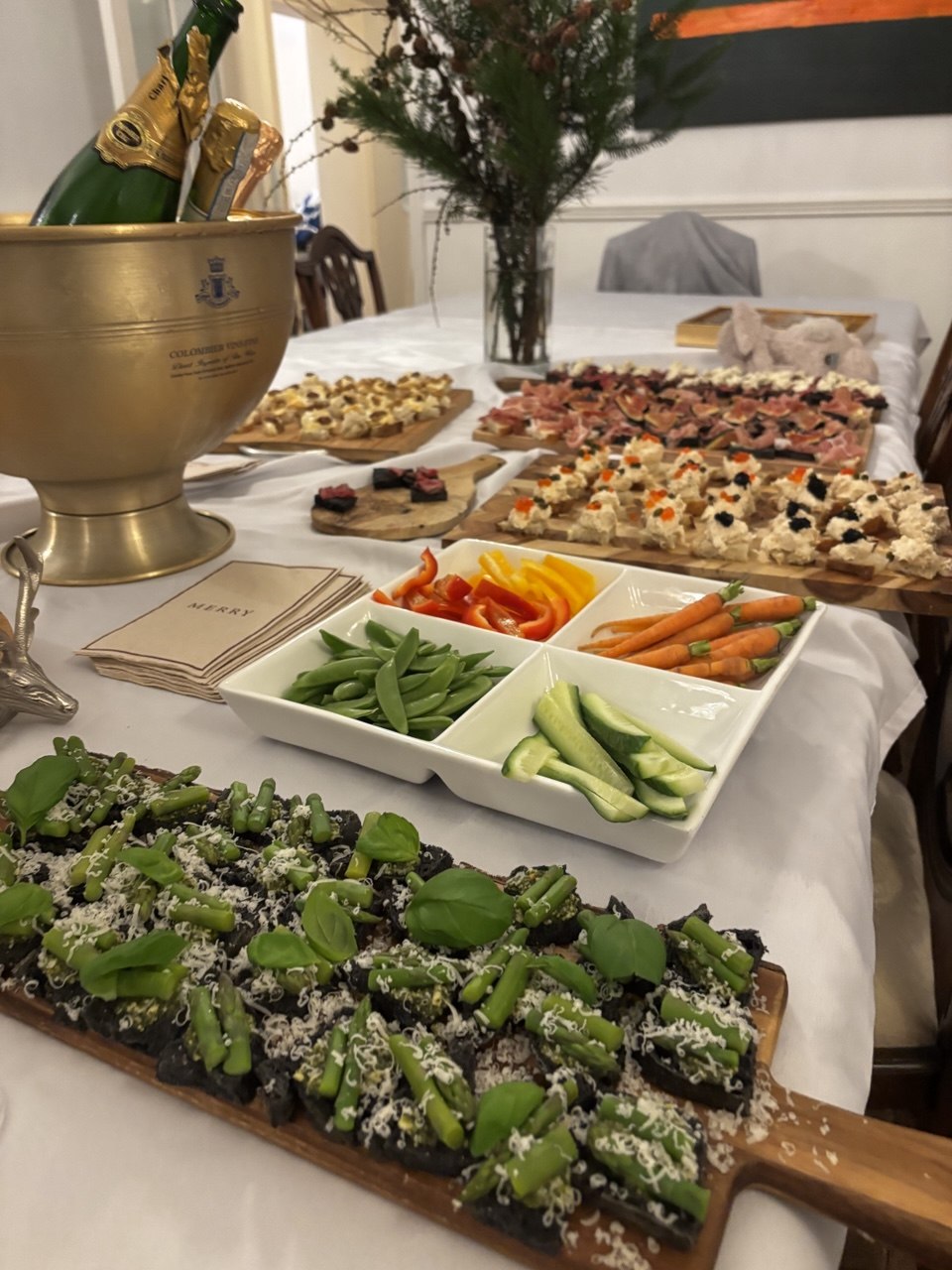 A long wooden serving board covered with dark rye bruschetta topped with roasted asparagus tips, grated Parmesan and fresh basil leaves, surrounded by other party dishes including a crudités platter, charcuterie boards and a gold champagne bucket in the background