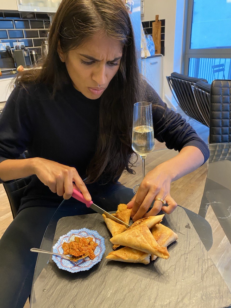 A woman with long dark hair frowns in concentration as she cuts into a golden samosa with a pink-handled knife. Several samosas are piled on a dark slate board and a small blue decorative bowl of spiced tomato chutney sits alongside. A flute of sparkling wine stands in the background on a glass dining table in a modern kitchen.