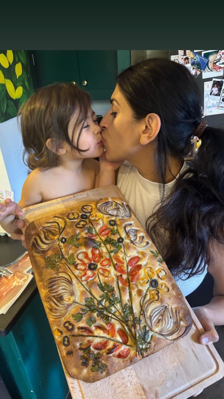 A woman with dark hair kisses a young toddler on the lips while both hold a large rectangular focaccia decorated with cherry tomatoes, red onion rings, black olive rings, yellow pepper strips and fresh parsley arranged to look like a garden of flowers and stems, displayed on a wooden board in a home kitchen.