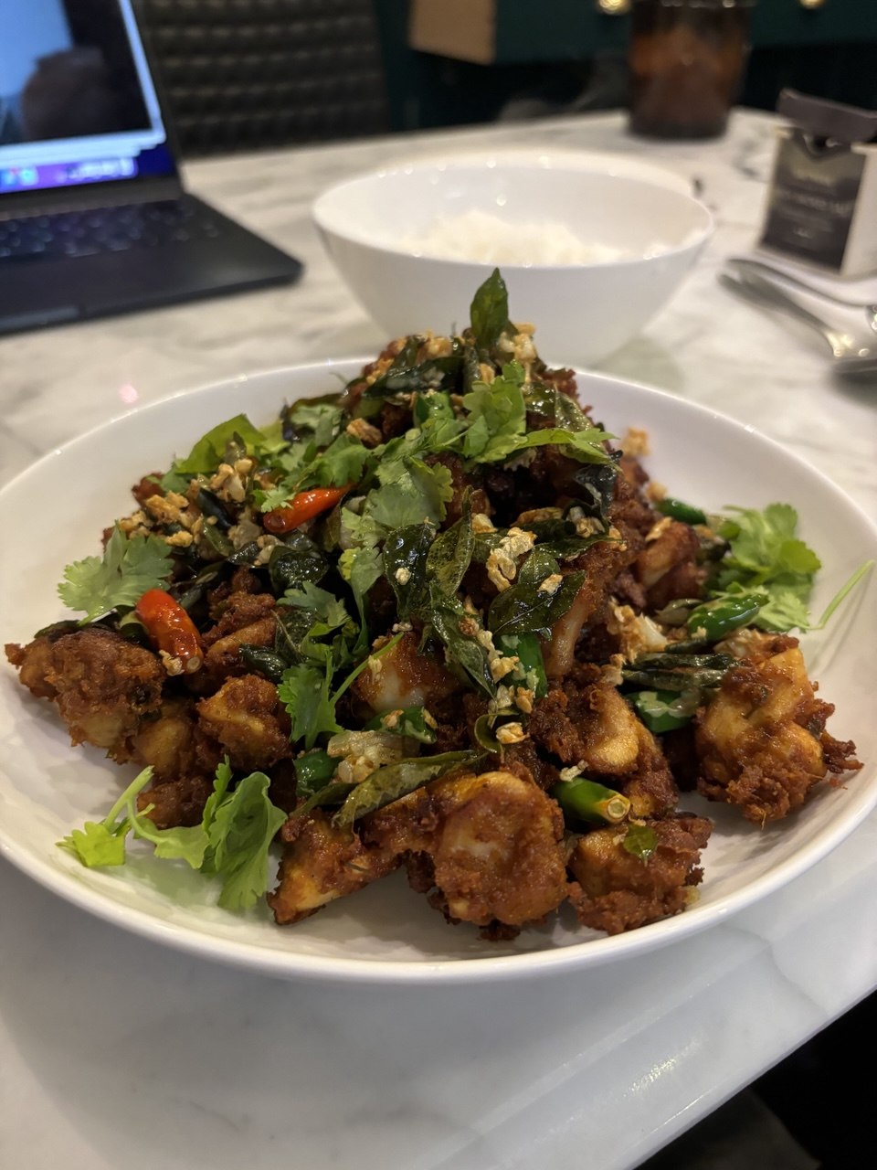 A white wide-rimmed bowl piled high with crispy fried chicken pieces tossed with fried curry leaves, green and red chillies, crushed garlic and scattered with fresh coriander, served on a marble restaurant table with a bowl of steamed white rice in the background