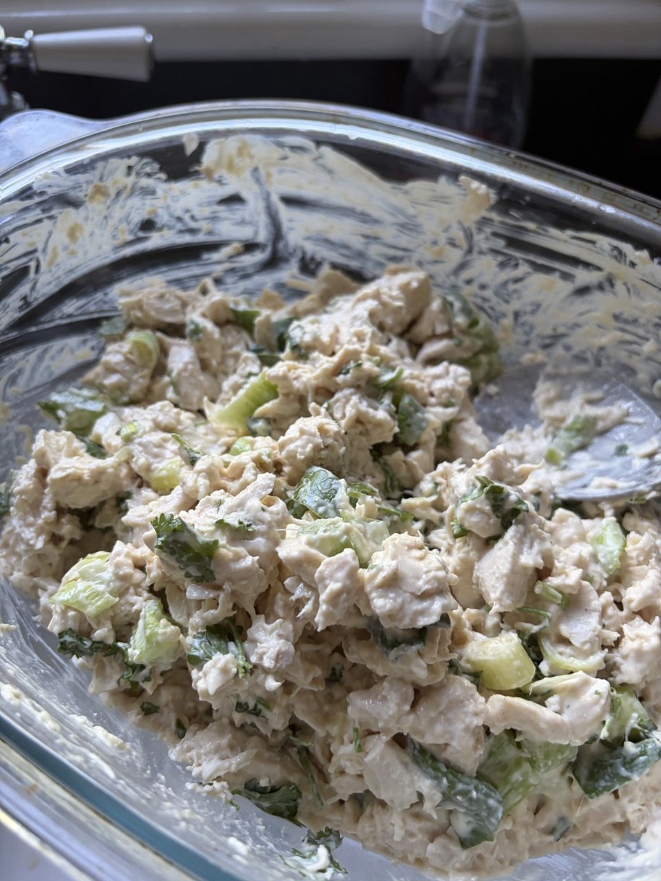 A large glass mixing bowl filled with creamy chicken salad made with chopped poached chicken, sliced celery and fresh green herbs, set near a kitchen sink.