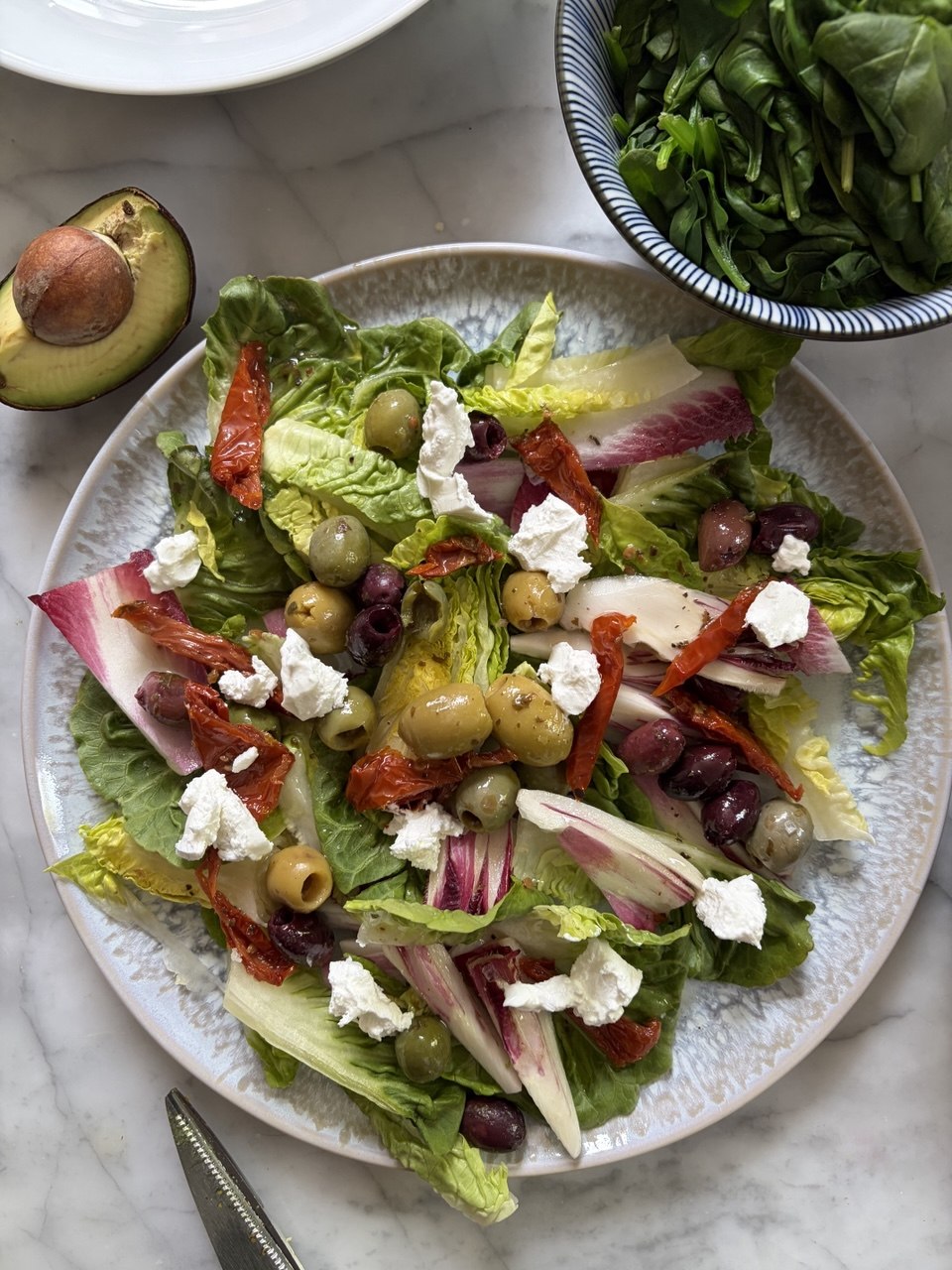 Overhead shot of a large white speckled plate holding a Mediterranean salad of romaine and chicory leaves topped with green and Kalamata olives, torn sun-dried tomatoes, crumbled goat's cheese and sliced avocado, with a bowl of spinach and a halved avocado visible alongside on a marble surface.