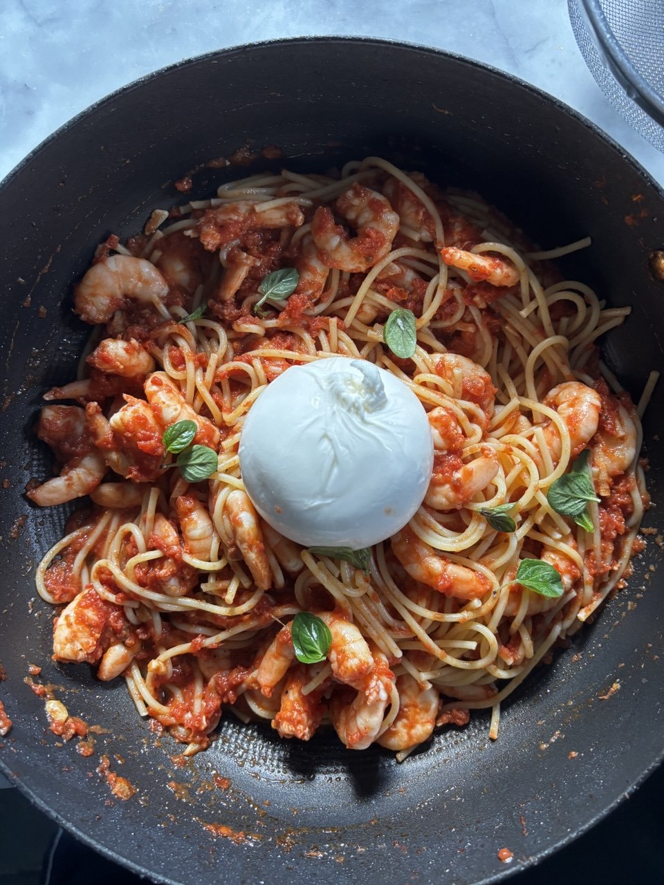 A large black wok viewed from above, filled with spaghetti in a rich red tomato and prawn sauce, scattered with fresh basil leaves and topped with a whole white burrata ball in the centre