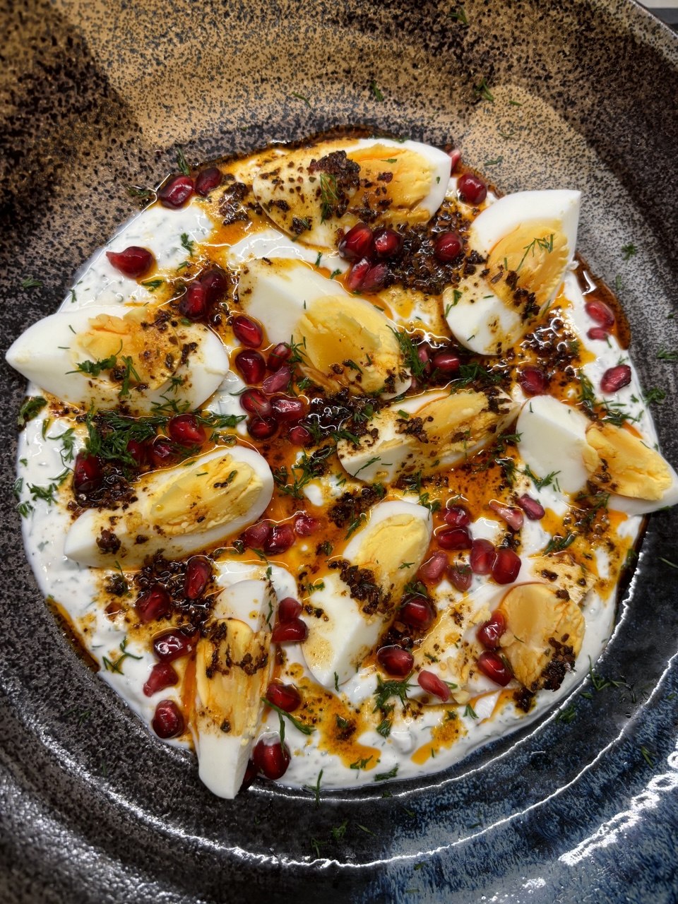 A dark speckled ceramic bowl filled with white skyr, topped with halved boiled eggs showing golden yolks, pomegranate seeds, fresh dill fronds and a drizzle of reddish-brown chilli butter.