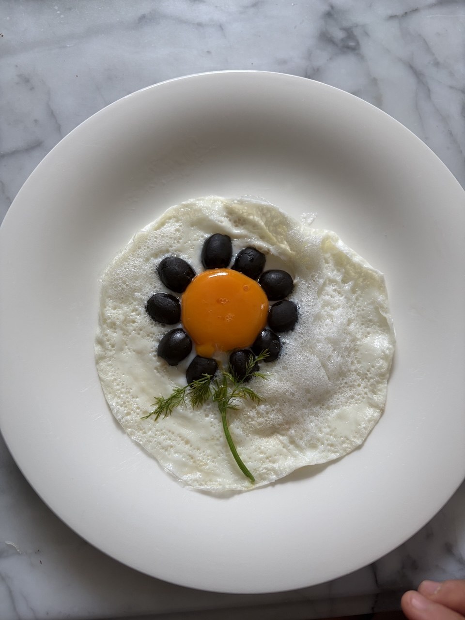 A fried egg on a white plate viewed from above. Black olives are arranged in a circle around the intact orange yolk to resemble flower petals, with two feathery dill sprigs placed below them to suggest a stem, all set against a white marble surface.