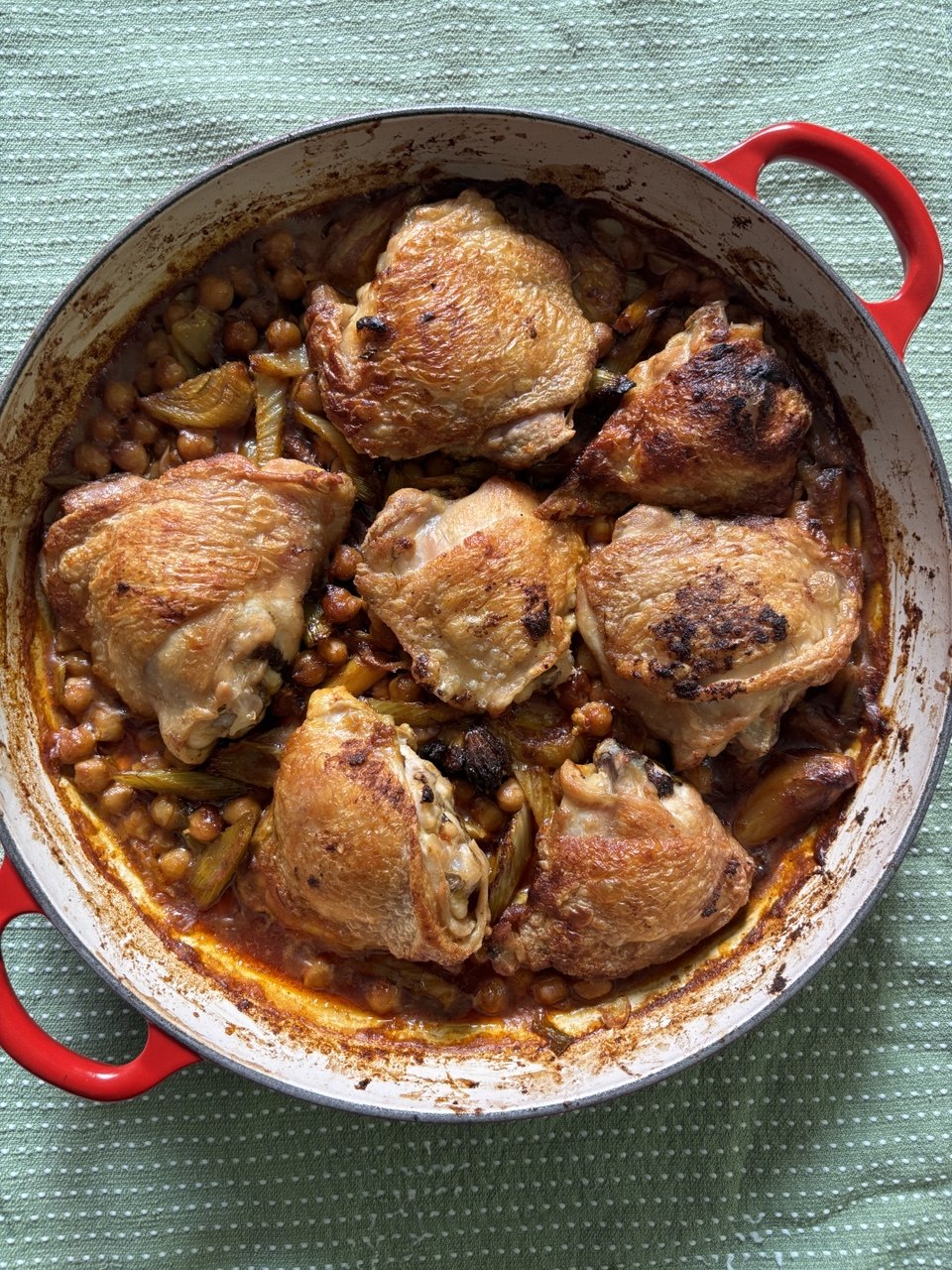Overhead view of a white and red enamel braising pan filled with six golden, crispy-skinned chicken thighs nestled among chickpeas, sliced fennel and a spiced reddish-orange sauce, set on a pale green linen cloth.