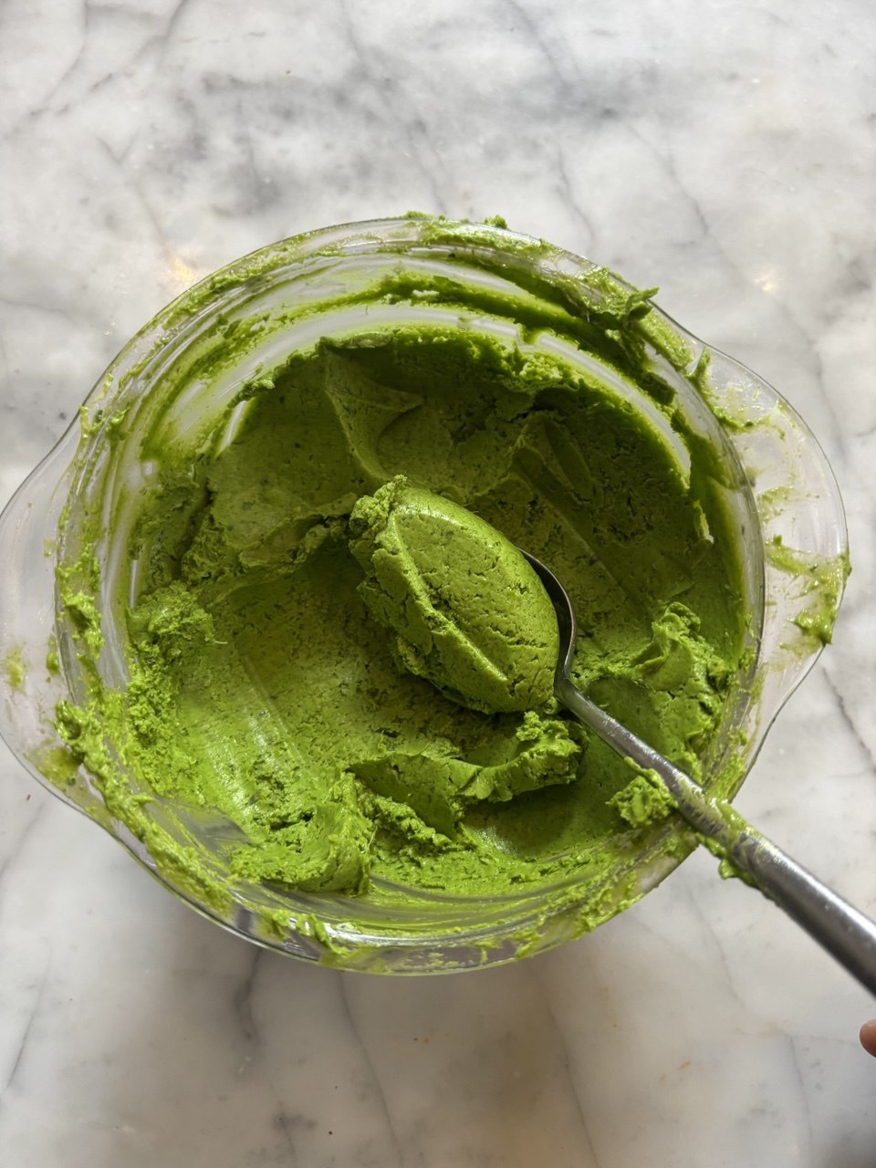 Overhead view of a glass blender cup filled with smooth, vivid green wild garlic butter, with a metal spoon lifting a generous scoop from the centre, set on a white marble surface.